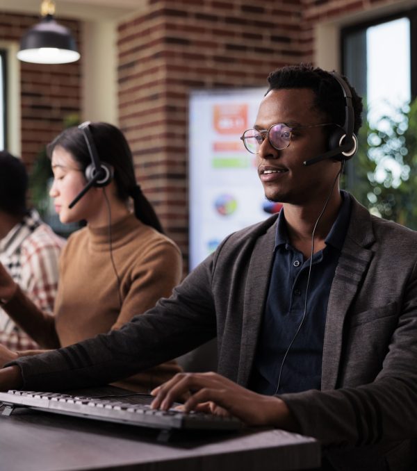 African american helpline employee working at call center reception with multiple monitors. Male operator using telecommunication to help clients at customer service support, remote network.